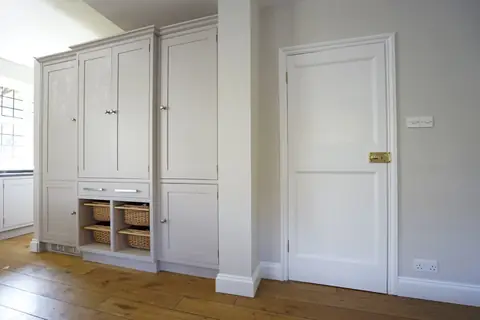 Tall grey painted larder cupboards with wicker basket drawers beside a white panelled door with a brass latch