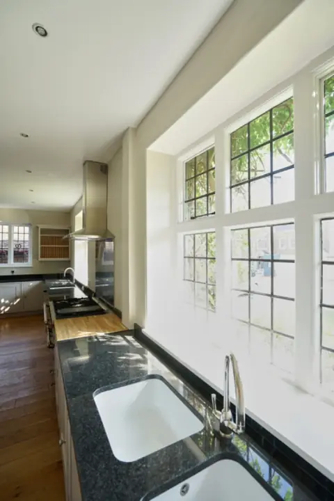 Dark worktop with twin white sinks under a wall of leaded windows, with the range and plate rack beyond