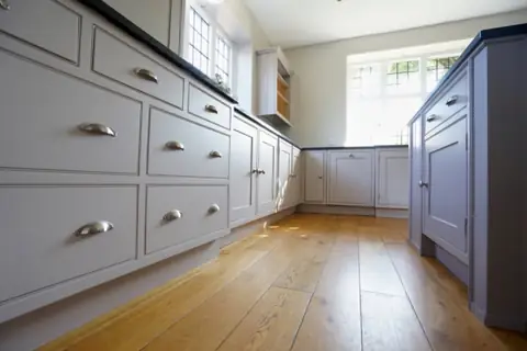 Low angle along grey painted drawers with cup handles towards a leaded window and plate rack