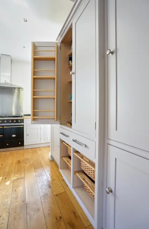 Tall grey painted larder with its door open showing wooden spice shelves, above wicker basket drawers