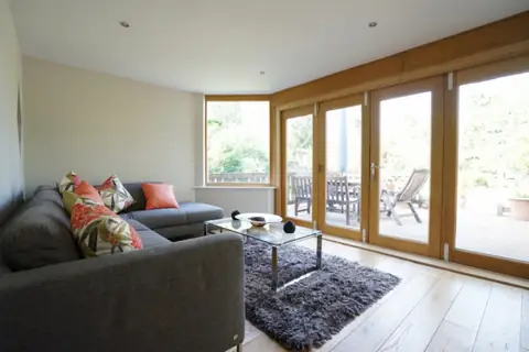 Family room with a grey sofa, glass coffee table and oak-framed folding doors open to a decked garden