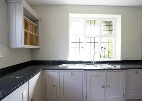 Grey handmade base cupboards and a dark stone worktop under a leaded window, with a painted plate rack on the wall