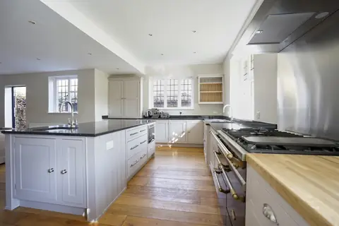 Open kitchen with a grey painted island, range cooker, tall cupboard, plate rack and leaded windows over oak boards