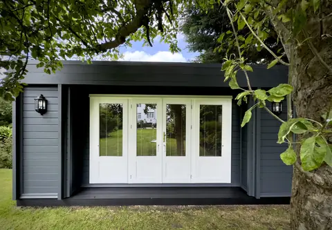 After: front of the garden cabin seen straight on, with dark grey cladding and white doors framed by a tree