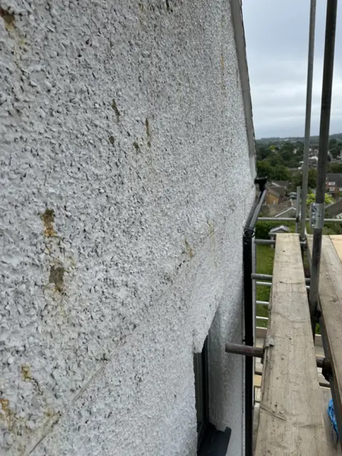 Close view along old pebbledash with brown rust spots and streaks above a window, scaffold board beside