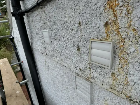 Heavy orange rust staining running through pebbledash around white wall vents beside a black downpipe