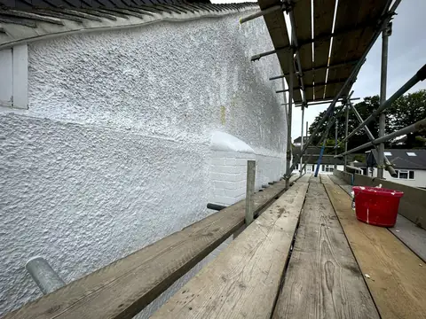 Long pebbledash wall being painted white seen from a scaffold walkway, with a red bucket on the boards