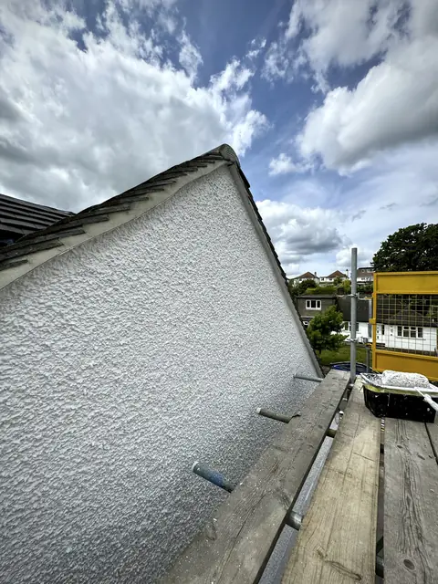 White painted pebbledash gable seen from the scaffold, a roller tray on the boards and houses on the hill behind