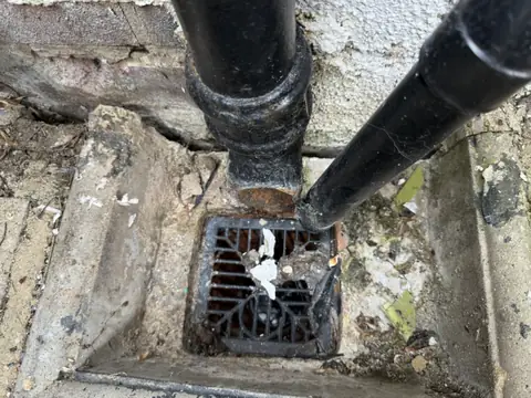 Dirty drain gully at the base of a wall with black downpipes, debris and old paint flakes around it