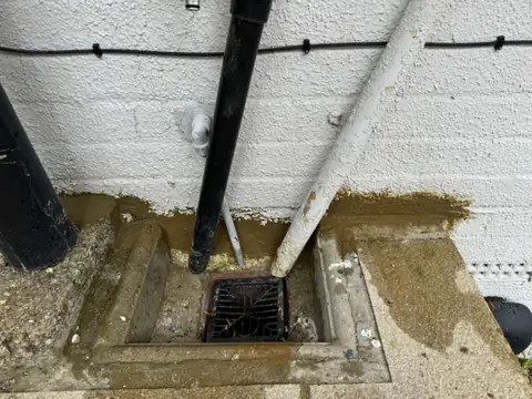 Drain gully below a white painted wall with pipes and a fresh sand-coloured repair along the base