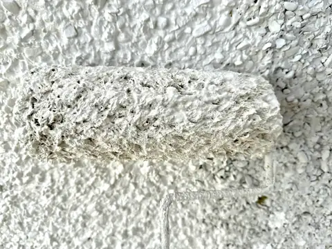 Close up of a thick pile roller loaded with white paint, held against freshly painted pebbledash