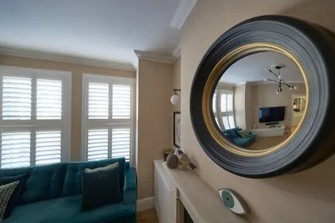 Convex mirror on a warm neutral chimney breast reflecting the lounge, with shuttered windows and teal sofa beside it
