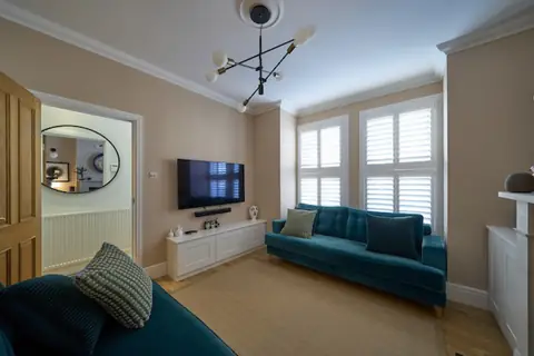 Lounge with oak door open to the hallway, wall-mounted TV, white cabinet and teal sofa under shuttered windows