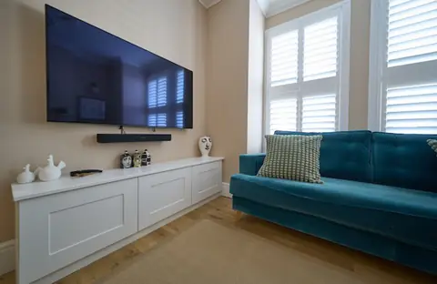 Close view of a lounge wall with TV and soundbar above a white cabinet, teal sofa and white shutters