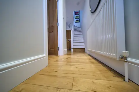 Low view along a hallway with oak floor, white skirting, long radiator and staircase at the far end