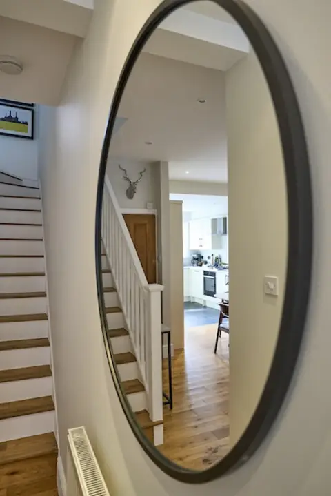 Round hallway mirror reflecting the staircase, white balustrade and kitchen beyond, with stairs to the left