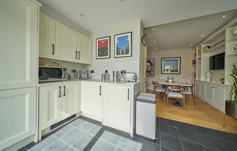 Kitchen corner with cream units and two framed prints, opening through to the dining area and shelving