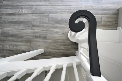 Dark painted handrail scroll seen from above, over white spindles and a wood-effect floor