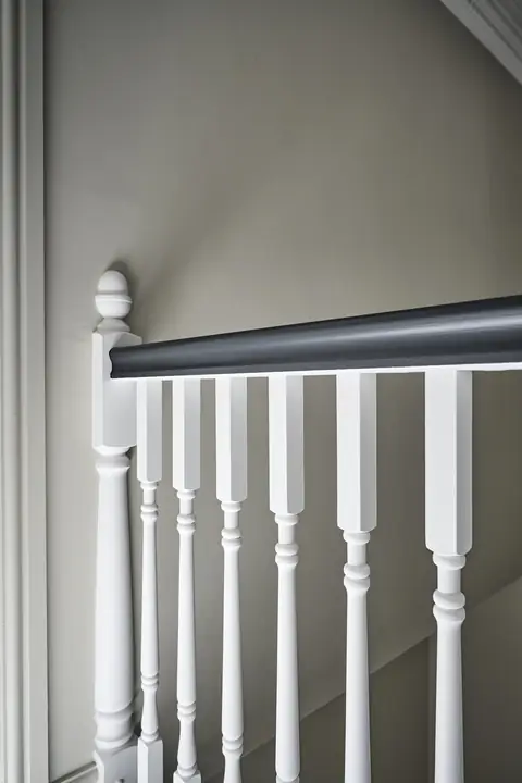 Close up of a dark painted handrail over turned white spindles and a newel finial, against a pale painted wall