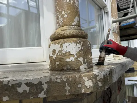 Gloved hand brushing a clear liquid onto a scraped-back stone window sill and pillar base