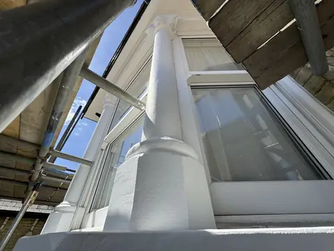 Looking up a smooth white painted bay window column to its carved capital, scaffold and blue sky above