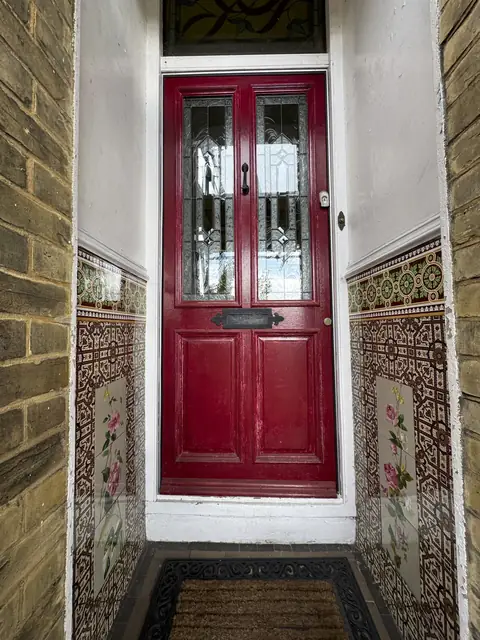 Faded red front door with leaded glass panels in a porch lined with Victorian floral wall tiles