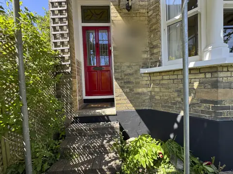 Red front door in a porch beside a white bay window, black painted plinth and brick steps in sunlight