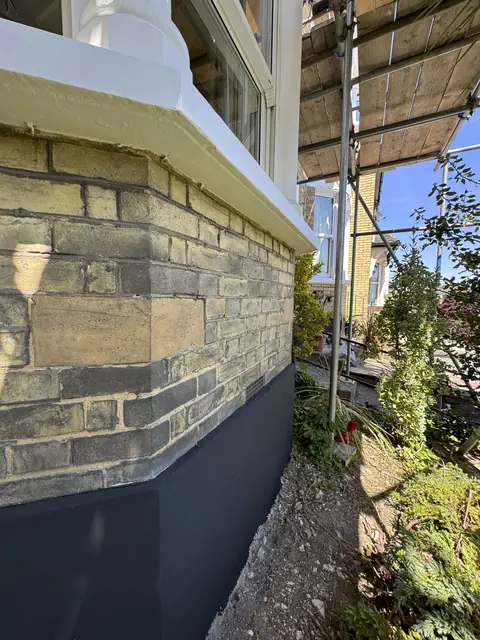 Side view of a bay with a white painted sill above yellow brick and a black painted plinth beside the garden