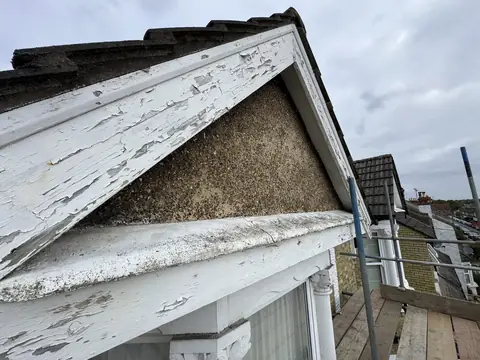 Pebbledash gable framed by white barge boards and fascia with paint cracking and peeling, seen from scaffold