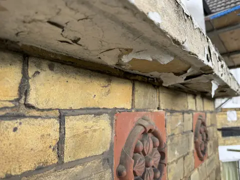 Underside of a stone bay sill with paint cracking and peeling, terracotta rosette tiles below