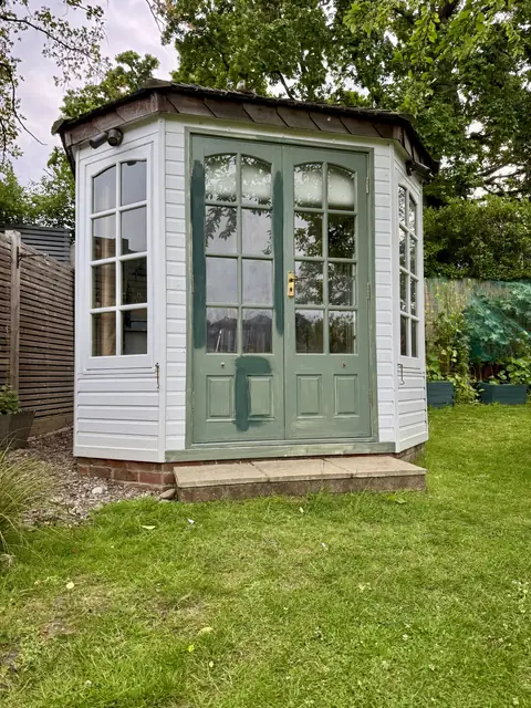 Octagonal garden office before work, white weatherboard, worn green double doors with darker paint patches, weathered roof