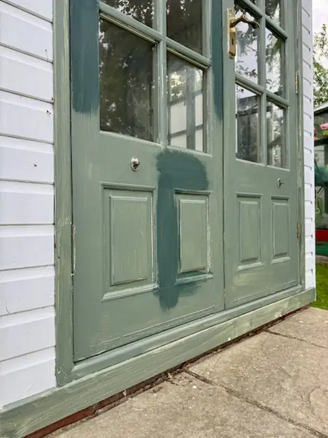 Lower panels of worn sage green garden office doors with darker green brushed patches and white weatherboard beside