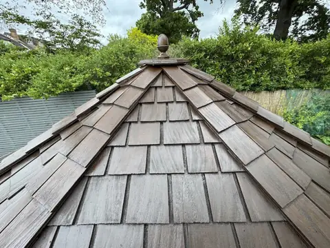 Cedar shingle roof stained rich brown with a new acorn finial on top, hedges and trees behind