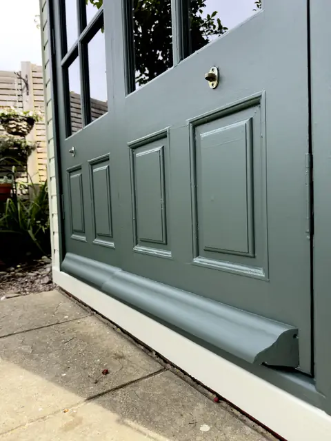 Dark green panelled garden office door with painted weather strip, paving slabs and potted plants beside