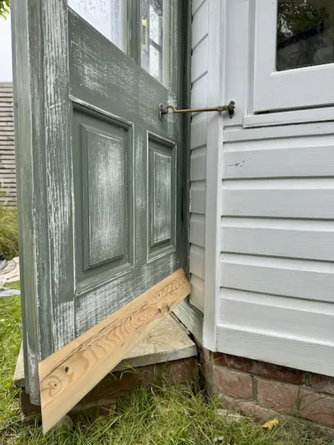 Open sanded garden office door with a bare timber weather strip held at its base and a brass cabin hook