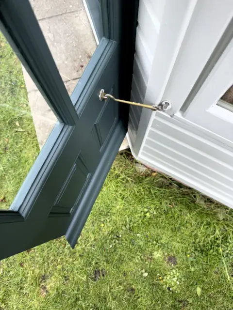 Dark green garden office door held open by a brass cabin hook against pale grey-green weatherboard