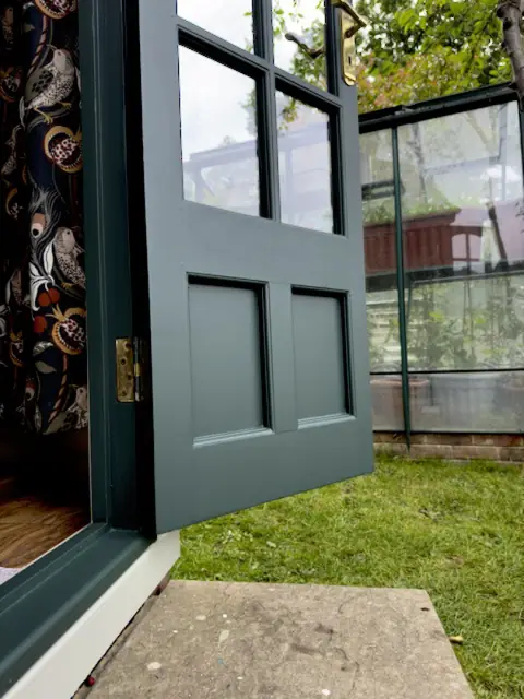 Open dark green garden office door with lower panels and step, a greenhouse on the lawn behind