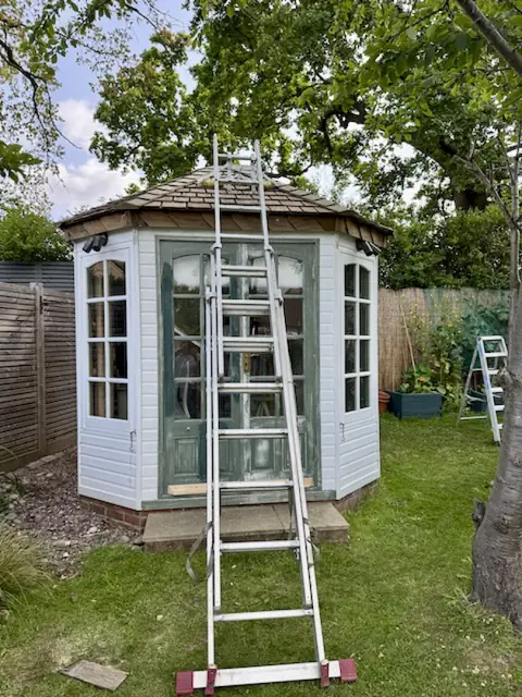 Extension ladder against an octagonal garden office with a step ladder on the lawn beside it