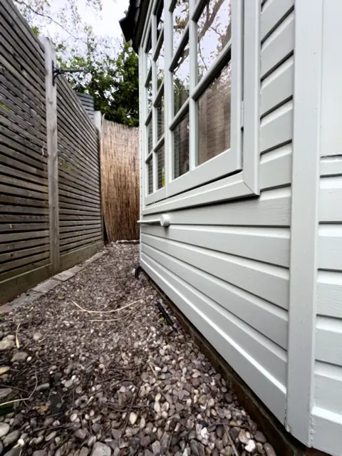 Side of the garden office in pale grey-green weatherboard beside a gravel path and slatted fence