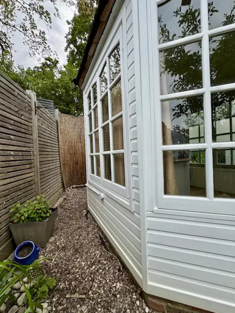 Side of the garden office in white weatherboard beside a gravel path, slatted fence and potted plant
