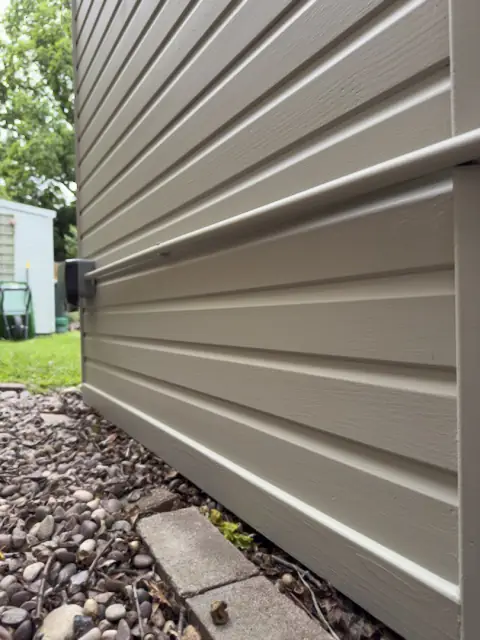Low angle along painted pale weatherboard with a conduit run and outdoor socket, lawn and shed beyond