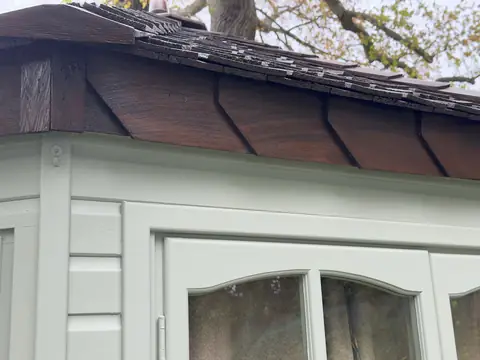 Dark brown stained eaves boards and shingle edge above a pale grey-green window frame