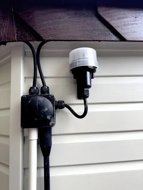 Black junction box and light sensor on pale painted weatherboard under dark brown eaves