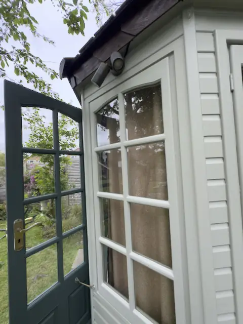Open dark green glazed door beside a pale grey-green window, twin spotlight under the eaves