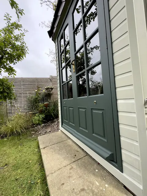 Dark green glazed garden office door and weather strip beside a paved step and planted border