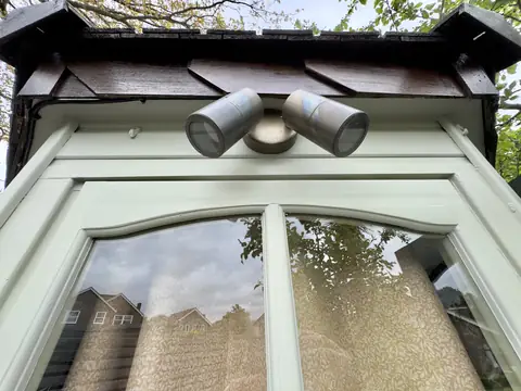 Twin outdoor spotlight under stained eaves above a pale grey-green arched window frame