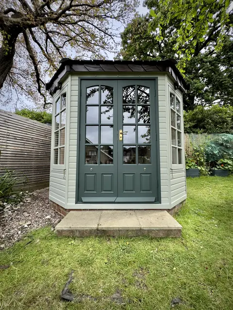 Octagonal garden office after restoration, pale grey-green weatherboard, dark green double doors and stained roof
