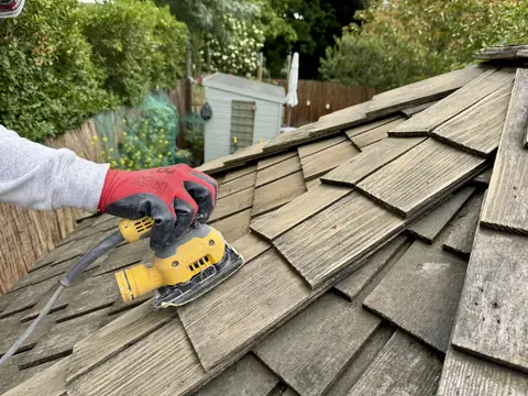 Gloved hand running an orbital sander over weathered cedar roof shingles, the moss already cleared