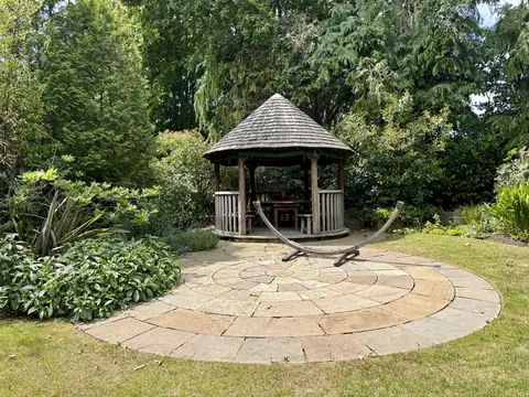Weathered grey timber circular gazebo with a shingle roof behind a round paved patio and hammock stand