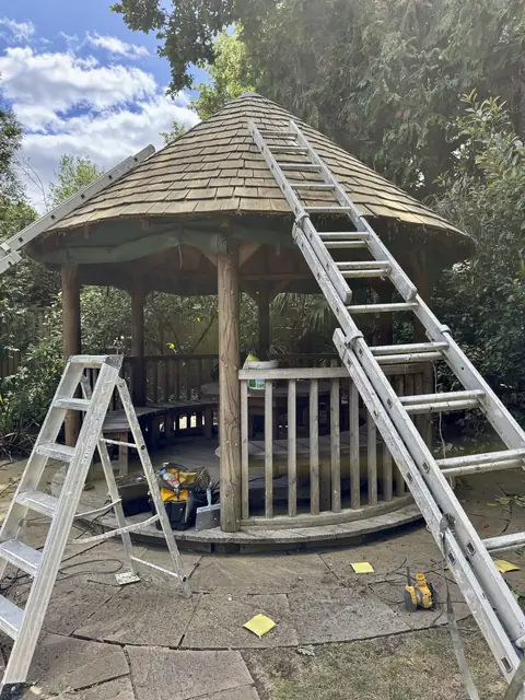 Extension ladder resting on a gazebo's shingle roof with a stepladder and tool bag on the patio
